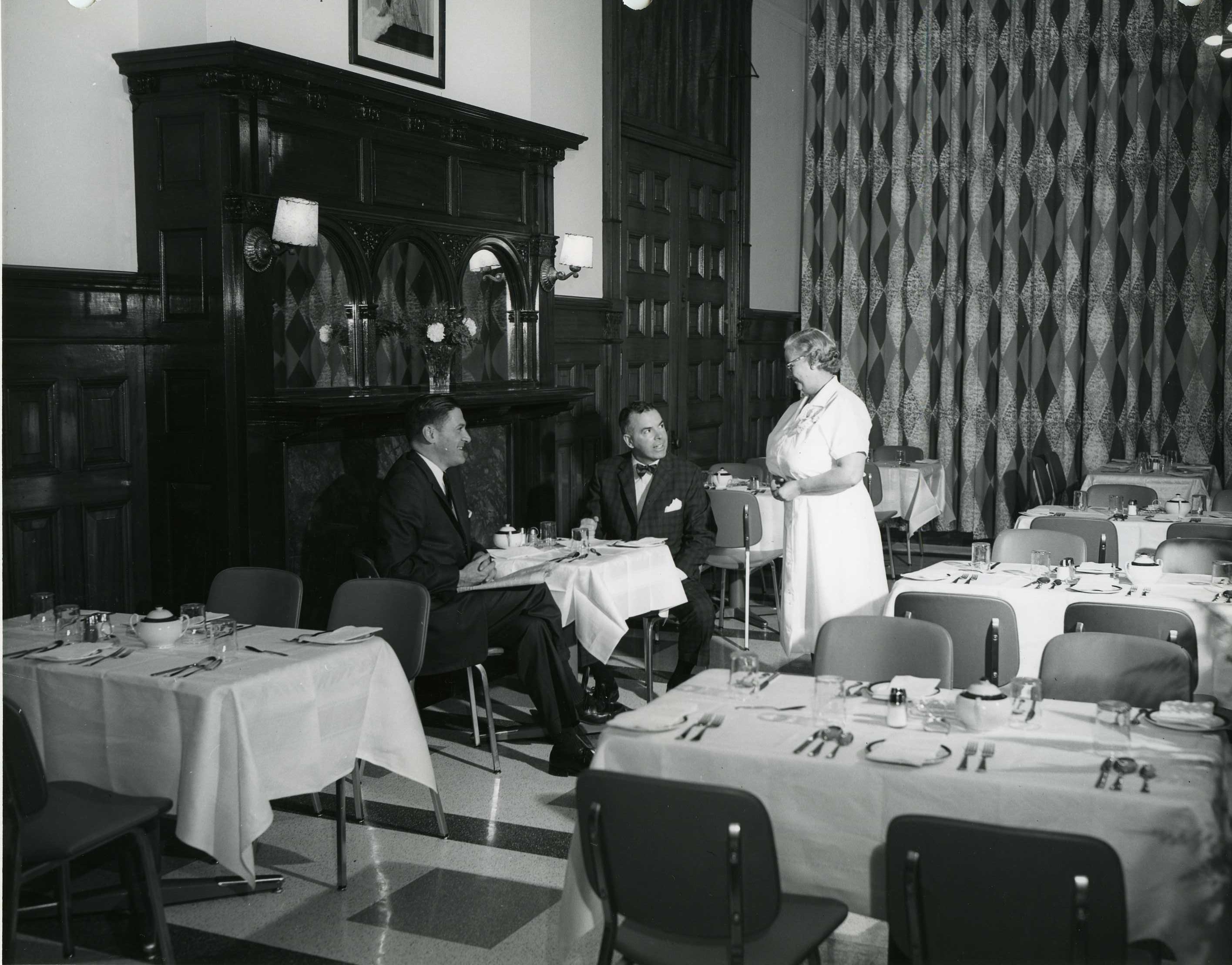 server standing next to two seated people at a table in a dining room