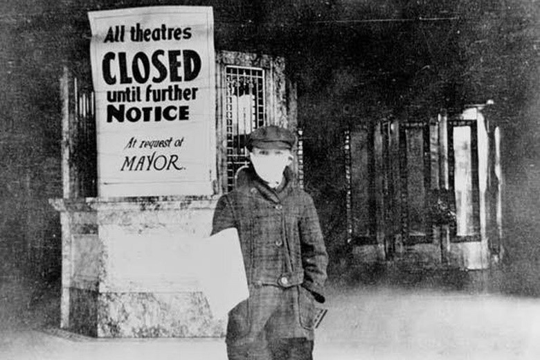 Photo showing a young newsboy in 1918 wearing a facemask and standing in front of a theatre box office which has a sign saying "All theatres closed until further notice at request of mayor."