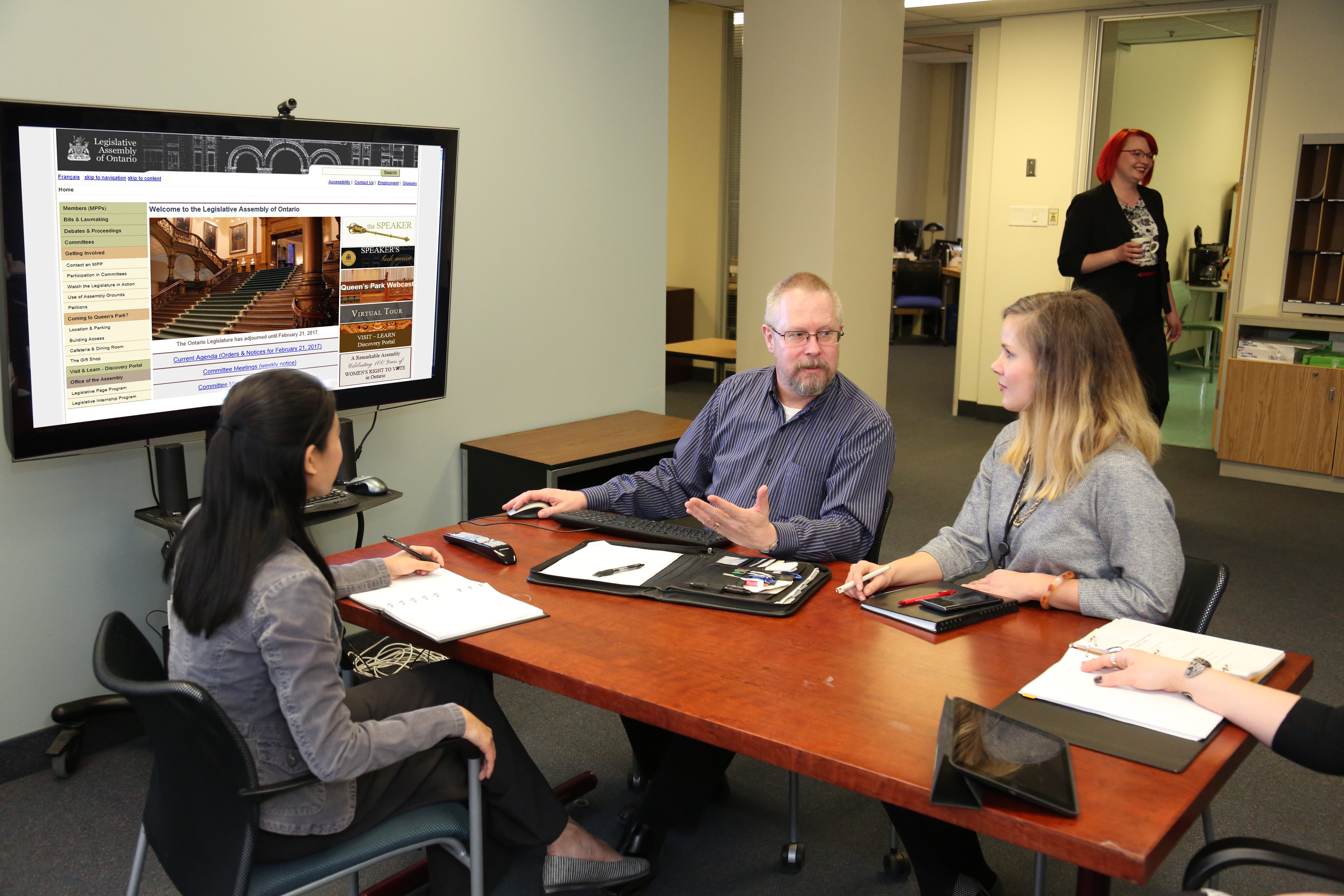 three people seated around a small table with a large screen in front 