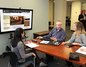 three people seated around a small table with a large screen in front